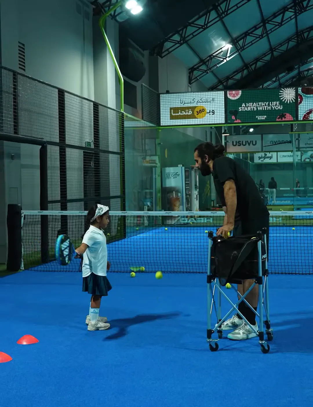 A coach with a young player and a ball cart on an indoor court at El Padel Academy