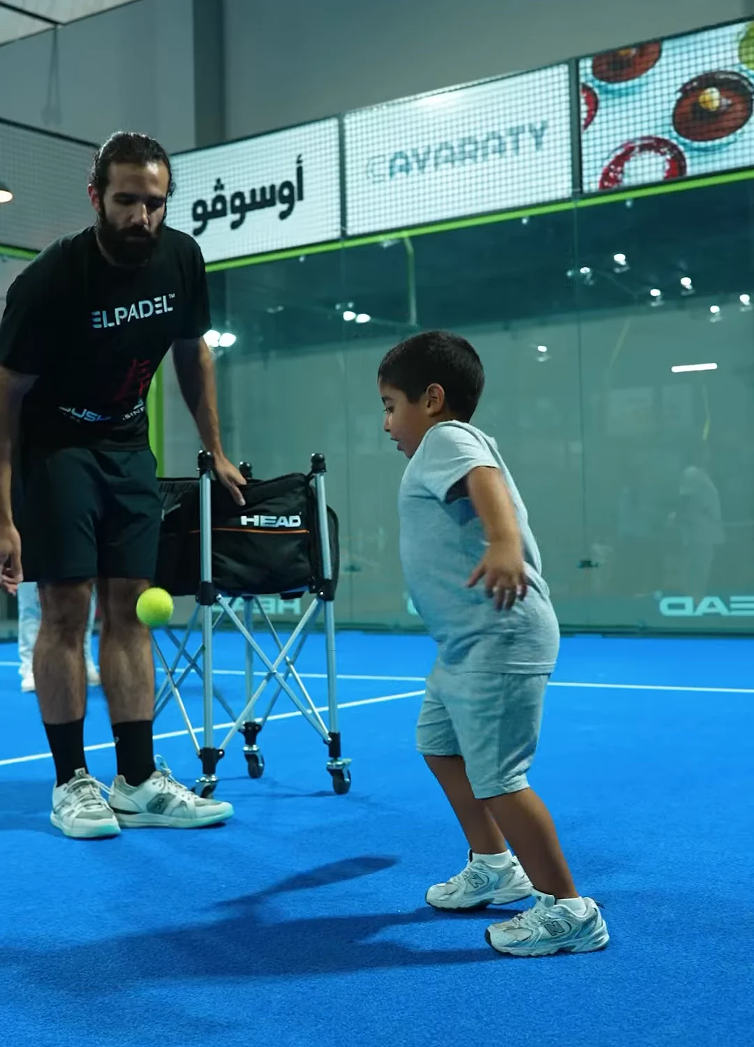 A coach in an El Padel shirt with a young player and a ball cart on court